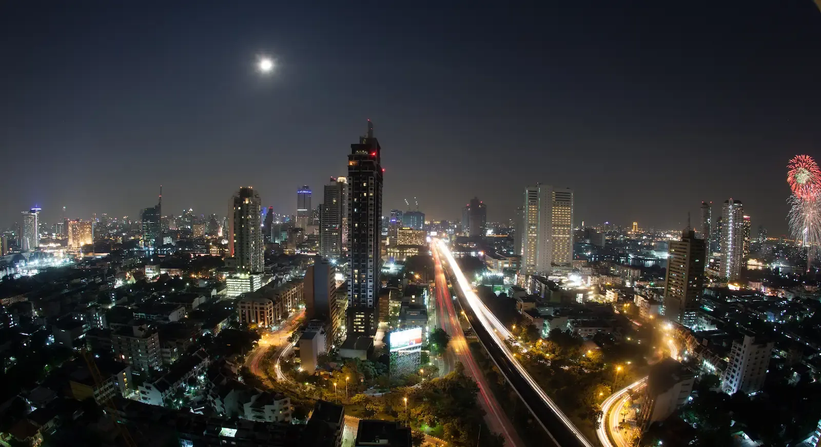 A nighttime cityscape capturing the vibrant lights of a city with tall skyscrapers, a clear moon, and fireworks in the background.